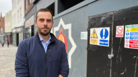 Luke Lutman, presidente de la Cámara de Comercio de Gloucester, se encuentra frente a una gran tienda vacía, que ahora está tapiada y pintada.