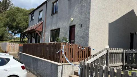 The exterior of a semi-detached property in Bonar Bridge. There is blue and white police taped across the front steps to the property.