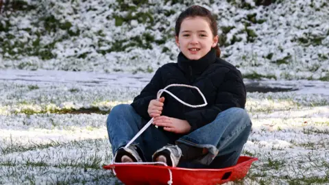 A girl rides down a hill in the snow in a park. Her sleigh is red. She's wearing jeans and a black jumper.