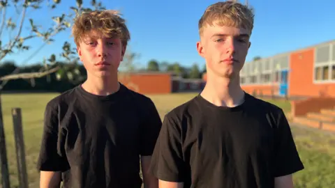 Two teenage boys, both wearing black t-shirts, looking into the camera with serious expressions. 