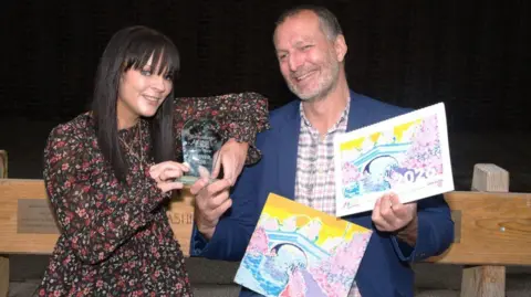 Lancashire County Council A middle aged man holding a calendar featuring a painting of a bridge accepts a glass trophy from a woman with long black hair wearing a floral patterned dress.