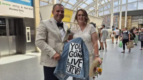 Gareth and Kim Gladman smile and hold up a blue denim jacket with the lyric "You and I are gonna live forever". He is wearing a cream white jacket and black bowtie while she is in a embroidered lace white wedding dress and holding a fan and bouquet of flowers. They are in the foyer of Manchester Victoria station with commuters in the background.