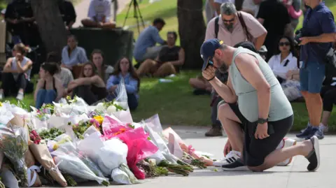 PA Media A man kneels in front of a large pile of flowers placed at Bondi Beach. Sitting on the grass around the pavilion, people are gathering.