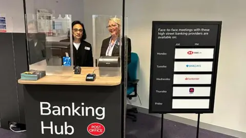 Mole Valley District Council Two people sat behind clear screens at a stand with a sign reading "Banking Hub".