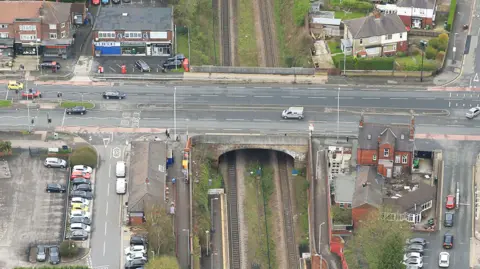 An aerial view of a road bridge over a railway line. Two vehicles can be seen on the road. Several low rise buildings alongside the road. Cars parked in a car park.