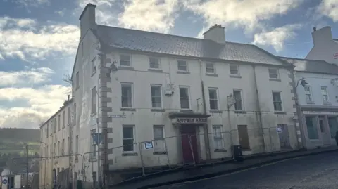 A white building with a red door and red lettering above the door which reads "Antrim Arms". The building is three stories high and is showing signs of wear. Two windows are boarded up and there is a plant growing out of the guttering. The building is surrounded by a metal railings