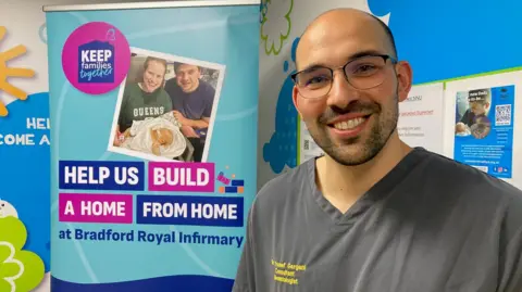 Charles Heslett/BBC A bald man in glasses and a grey hospital tunic standing next to a poster with the fundraising details on it 