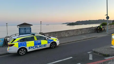 A police car is parked at the side of the road. The road overlooks Plymouth Sound with Drakes Island in the background. The photo has been taken as the sun is setting.
