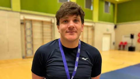 Callum Perry has thick dark, short hair. He is wearing a black t-shirt and has a purple "DBS" lanyard around his neck. He is standing in a sports hall.