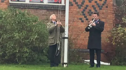 Niall Roberts A man in a dark suit and coat, playing a bugle, alongside a man lowering a flag on a white flag pole. Behind them is a red brick building with darker bricks marking out a diamond pattern in the brick work with green bushes in front of the building