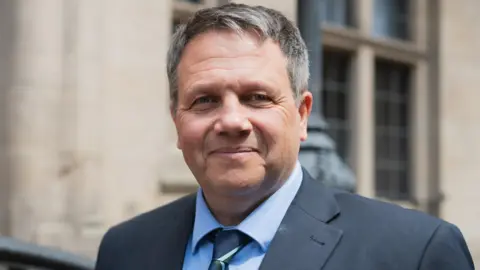 Staffordshire County Council A man with grey hair is wearing a tie, shirt and blue jacket and is prominent in the image. The background, a building, is blurred. 