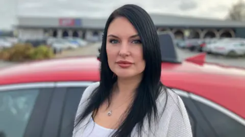 A woman with long dark hair is stood alongside the side of a red driving instructor's car. She is looking directly at the camera