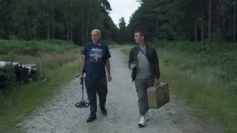 Nub Films & Wyrd Kingdom Pictures Two men, one on the left holding a metal detector and the one on the right with a square shaped bag, are walking down a pathway in the middle of a forest.