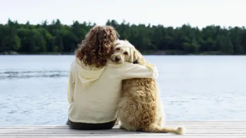 Getty Images A woman sits on a jetty next to the water with her right arm around the neck of a sandy-coloured dog. The dog has its head turned to the camera.
