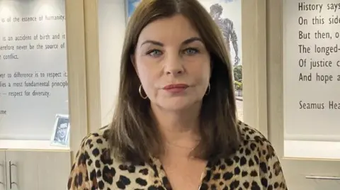 A woman has long brown hair and is wearing a leopard print top with hoop earrings. She is standing in front of walls with writing on them.