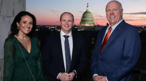 Three people in formal attire pose for a picture at sunset with Capitol Hill visible in the background. The woman on the left has black hair and is wearing a purse which says 'America 250'. The man in the middle is wearing a dark suit, the man to his right a blue suit.