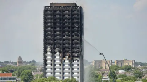 A smouldering burnt-out high-rise building in the foreground of a view of London. The building is being hosed by a fire appliance attached to a cherry picker. 