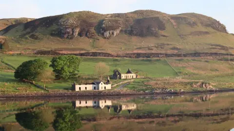 Michelle Popp Two small houses at the bottom of a tall grassy hill. The house reflects onto the very still water. It appears to be the end of the day, and the light is fading.