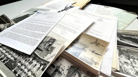 Old black and white photographs and a number of typed documents spread across a table. The photos are of servicemen and women, military ships and planes. 