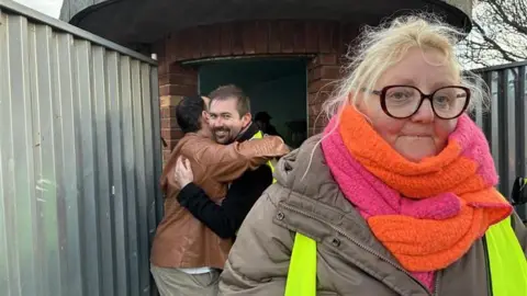 Robby West/BBC Liz Withington, a woman who is standing at the bus shelter looking directly at the camera. She is wearing a brown coat with a fluorescent hi-vis vest over the top and a pink and orange scarf.