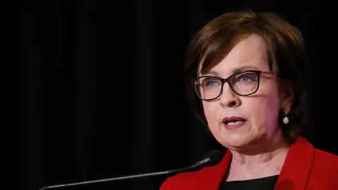 Pacemaker A woman with dark brown hair is talking at a podium. She is wearing a red blazer and dark rimmed glasses. 