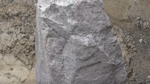 Rough, grey stone slab standing upright in a patch of bare, light-brown dirt. The stone surface is uneven and weathered, with small pitted marks and faint cracks - almost looking like a face. Loose soil and shallow grooves surround the base.