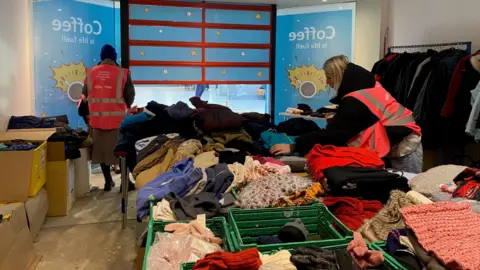 Two women wearing pink high-visibility vests organising crates of winter clothing in an industrial unit. The shutters at the entrance to the unit are half-closed and on one side there's a clothes rail with a row of coats on it.