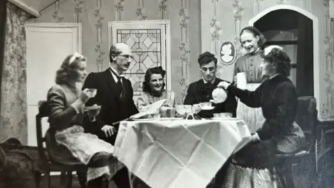 SMHWCC A black and white photograph of a stage set with five people and a maid round a table, with one actor serving tea from a pot