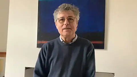 Peter is standing in a living room with a painting behind him of a night sky. He is wearing a blue pullover and glasses