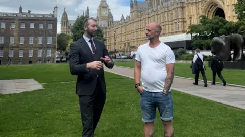 Will Stone and Adam Parker stand on College Green outside the Houses of Parliament. Stone wears a dark suit, shirt and tie. Parker wears denim shorts and a white T-shirt. 