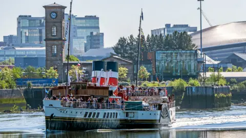 The Waverley sailing near a harbour that has a lot of industrial buildings and a tower with a clock. The vessel has lots of miniature multicoloured flags and passengers on board. 