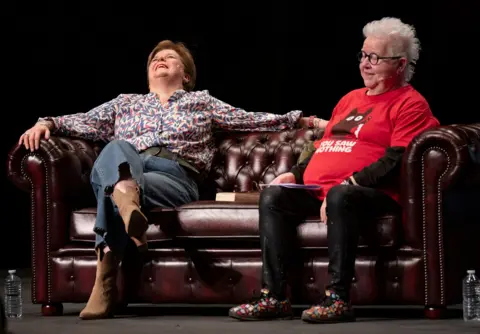 PA Media Nicola Sturgeon sitting on a leather chesterfield beside Val McDermid. Sturgeon is rocking back in the chair as she laughs