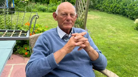 Roger Nightingale sits on a wicker chair in his garden with his hands crossed together. He is wearing a blue jumper and blue checked shirt underneath.