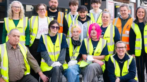 Volunteer It Yourself A group of volunteers outside Voodoo Daddy's in Norwich, wearing fluorescent hi-vis vests. The group is made up of 13 people in the picture.
