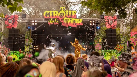 Isle of Wight Festival - Callum Baker Two people dressed in a gold and a silver mirror suit stand on stage at Cirque de la Quirk in front of an audience all wearing fancy dress.