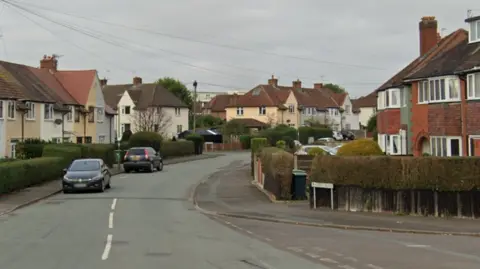 Google A residential street with cars parked on either side. A junction of the road can be seen heading off to the bottom right of the photo. There is a dotted white line up the centre of part of the road but it disappears further down. The houses on one side are flat-fronted with different colours to the walls including light blue and cream while the homes opposite are mainly red brick with bay windows.