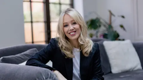 Emma Kay is seen sitting on a grey sofa with white cushions. She has long, wavy blonde hair and is wearing a navy suit jacket and a white t-shirt.