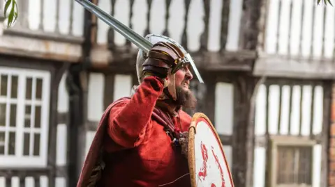 A man dressed in a Viking outfit, including a red shirt and cloak, as well as a helmet, sword and shield, stands side-on in front of a white and black building.