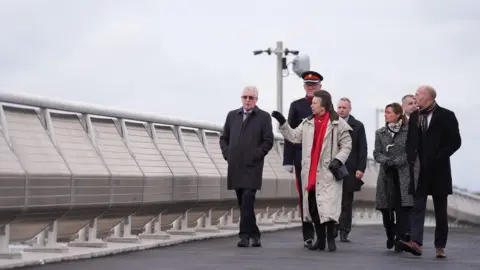 PA Media Princess Anne fronts a small group of people as they walk along Lowestoft's new Gull Wing bridge. She is talking to a gentleman next to her and using her hand to demonstrate an action.