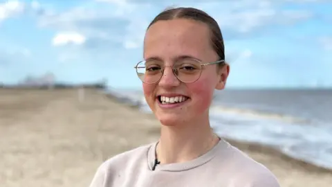 Mary Catchpole, smiling with her hair tied back and wearing glasses. She is standing on a beach near her home in Norfolk