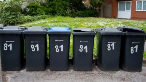 A row of six black, numbered wheelie bins on a pavement in front of a grass lawn and a house