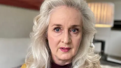A close-up of Nicola Bannon, a woman with shoulder-length wavy white hair. She is sitting on a sofa wearing a mustard cardigan over a black top. 
