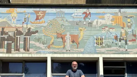 The pixel-like mosaic mural in muted blues, yellows. greens and browns shows key moments and figures from the county's history, including he Venerable Bede and the Viking raid on Lindisfarne (793 CE), the founding of Durham Cathedral (1093), the Penshaw Monument (1844), Captain James Cook (1728-79), the county’s collieries and the steel works at Redcar. Bennett Zon is standing beneath the artwork. He has white hair and beard and wears glasses.