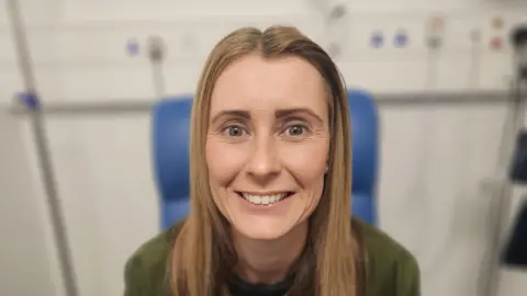 BBC Natalie is smiling at the camera, she has blonde shoulder length hair and is wearing a green jumper. She is sitting in a blue hospital chair.