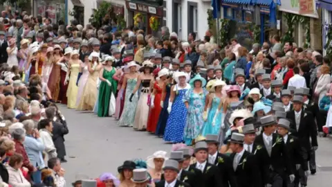 Formally dressed dancers line a street in Helston as part of the celebration. Onlookers stand either side of the street and watch performers. 