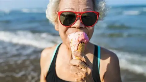 Age UK An older woman with short, white, curly hair in a black bathing suit is eating an ice-cream by the sea. She is wearing red-framed sunglasses. The ice-cream is pink and it is dripping down a waffle cone. The woman's mouth can't be seen behind the ice-cream but the rest of her face would suggest that she is smiling.