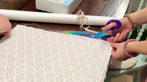 Remnant Revolution A hand is holding scissors and cutting alongside some fabric laid out on a wooden table.