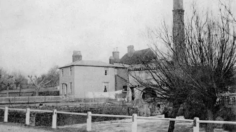 A black and white image of a mill next to a stream.