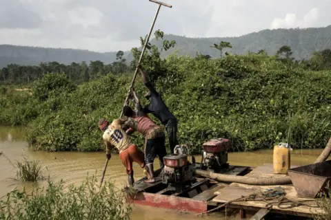 Getty Images Tiga pendulang emas ilegal berdiri di atas pompa bermotor untuk mengikis dasar sungai saat mereka mencari setitik emas di daerah Kibi di Ghana selatan pada 12 April 2017. Sungai itu berwarna kuning kecokelatan, dengan semak-semak lebat di kedua sisinya.