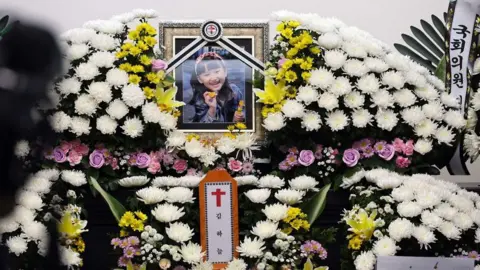 EPA-EFE/REX/Shutterstock A framed portrait of Kim Ha-neul, smiling brightly with a pink hairband, mounted on a pile of white chrysanthemums.